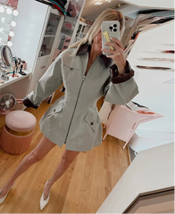 Person wearing a light gray coat taking a mirror selfie in a room with a vanity table.