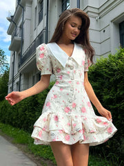 White mini dress with a white pointed collar, short puff sleeves, pearl button front, and a tiered ruffled hem.