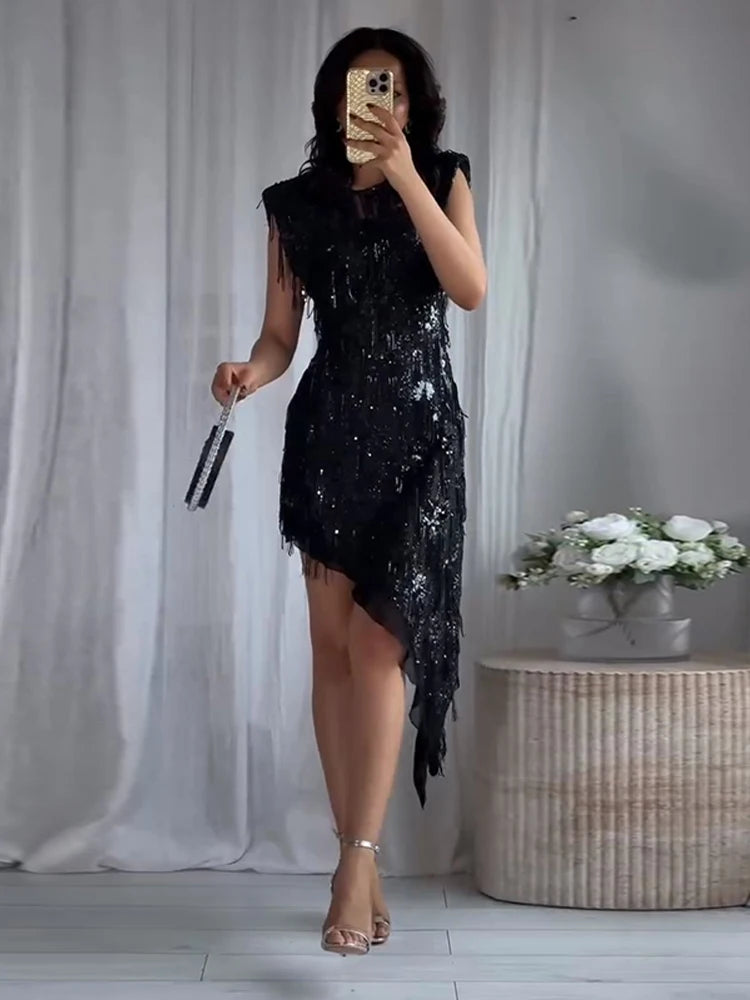 Person wearing a black sequin dress taking a mirror selfie in a room with white curtains and a woven basket.