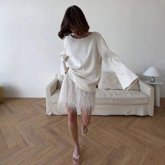 Woman in a white sweater and sheer skirt standing in a minimalistic room with a sofa and lamp.