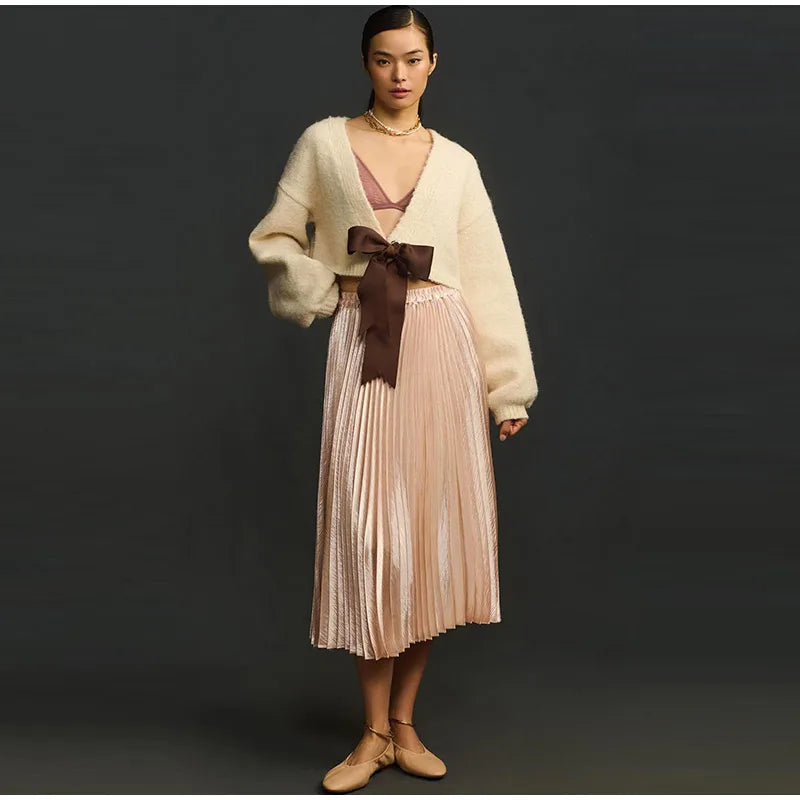 Woman wearing a cream cardigan, pink pleated skirt, and brown shoes on a black background