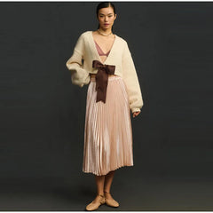 Woman wearing a cream cardigan, pink pleated skirt, and brown shoes on a black background