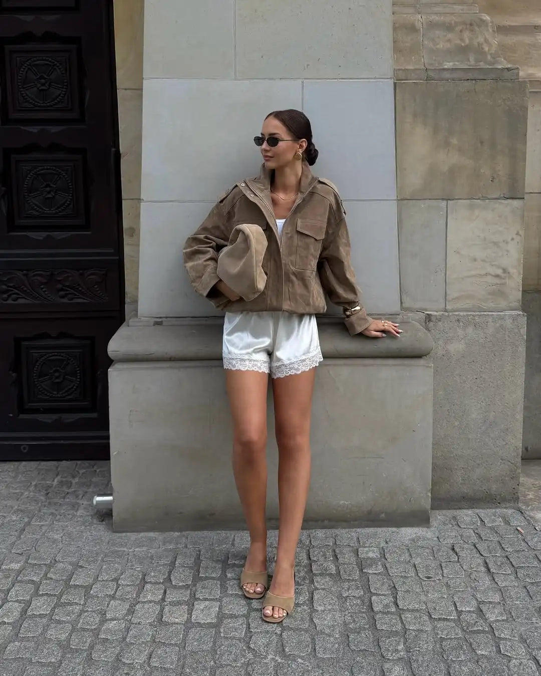Woman in a brown jacket and white shorts standing against a stone wall.