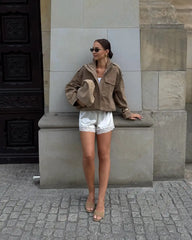 Woman in a brown jacket and white shorts standing against a stone wall.