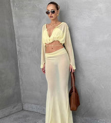 Woman in a light yellow outfit holding a brown handbag against a gray wall.