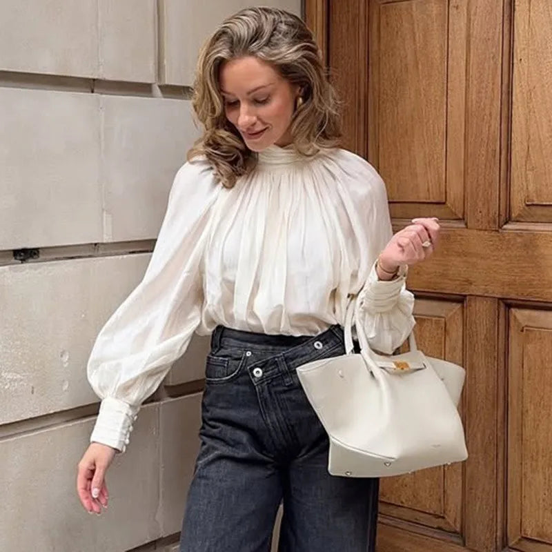 Woman in a white blouse and blue jeans holding a white handbag indoors.