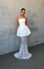 Woman wearing a strapless white dress with sheer overlay against a plain wall.