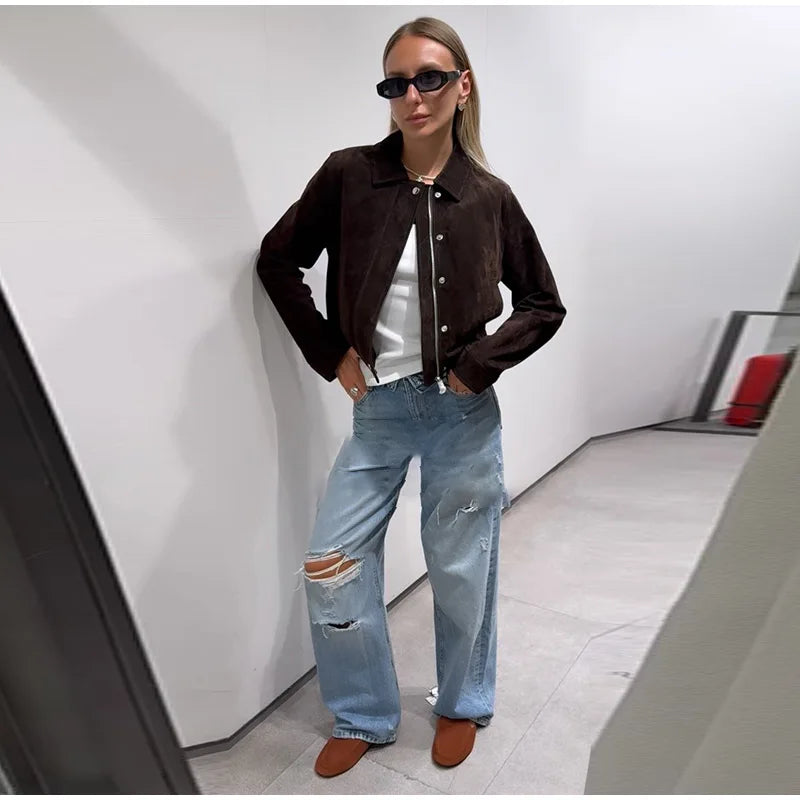 Person wearing a brown jacket, white shirt, and ripped blue jeans in a modern interior setting.