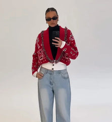 Person wearing a red patterned cardigan, black turtleneck, white shirt, and light blue jeans against a plain background.