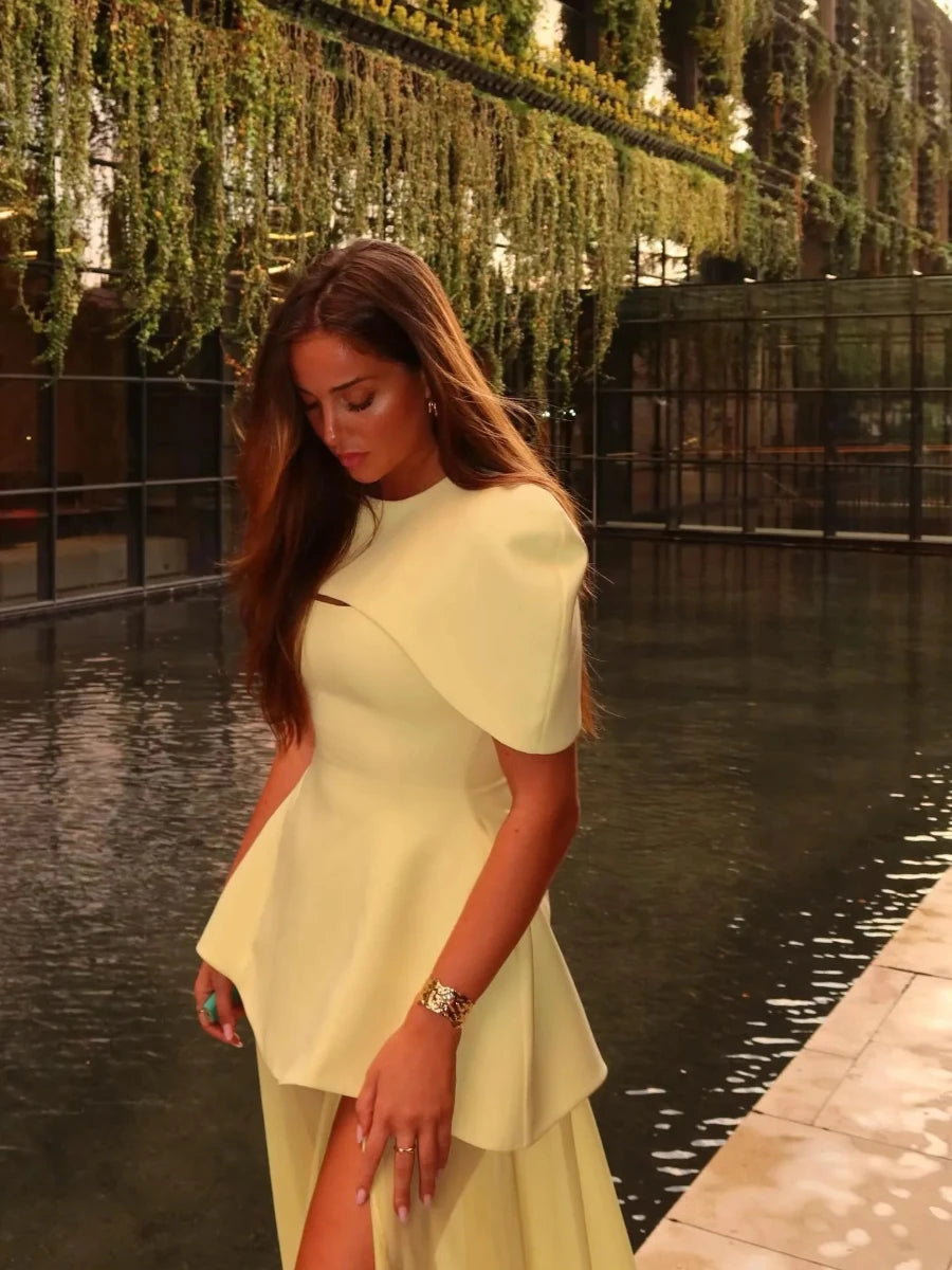 Woman in a light yellow dress standing by a water body with greenery in the background
