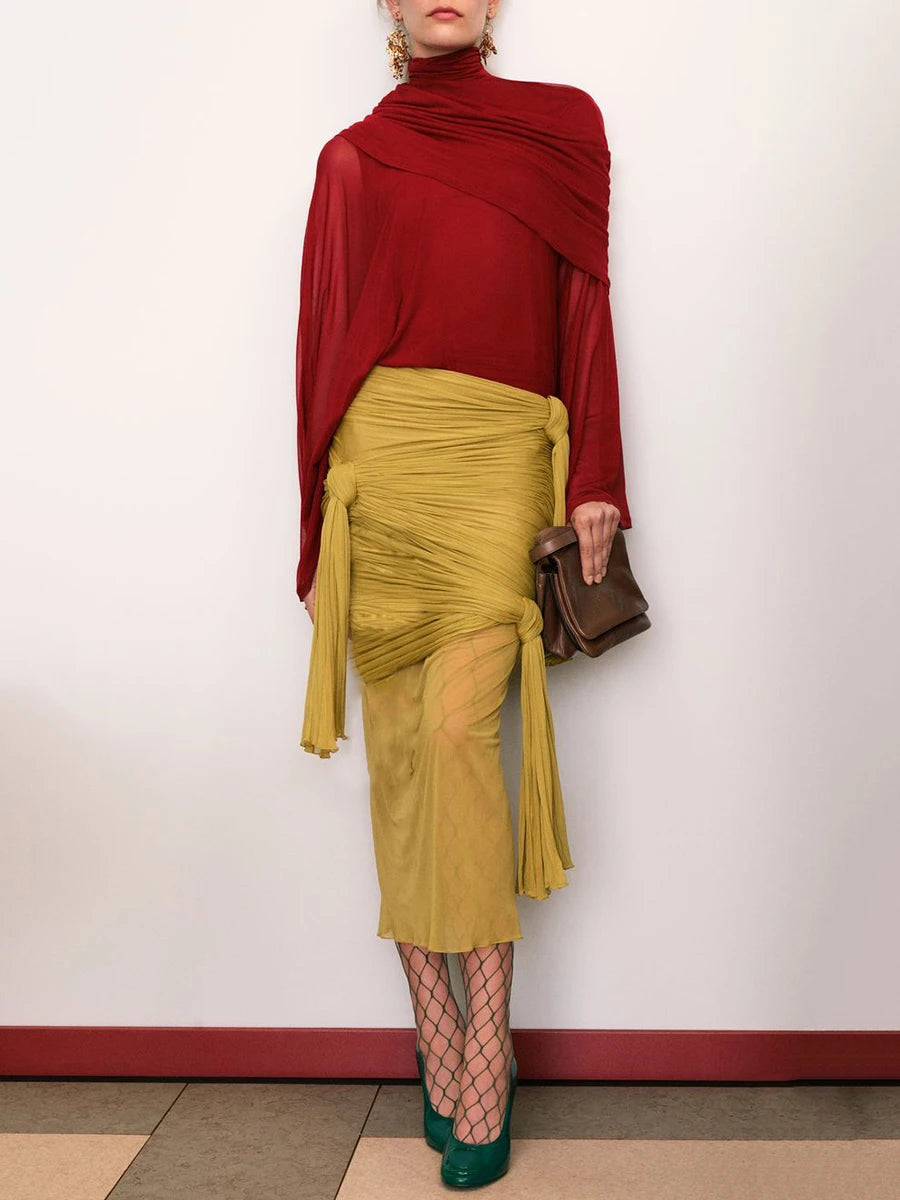 Person wearing a red top and yellow skirt with fishnet stockings and green shoes against a white wall.