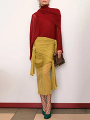 Person wearing a red top and yellow skirt with fishnet stockings and green shoes against a white wall.