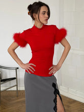 Woman wearing a red top with feather sleeves and a gray skirt in an indoor setting.