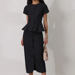 Woman wearing a black polka dot outfit with a peplum top and wide-leg pants, holding a beige handbag against a white background.