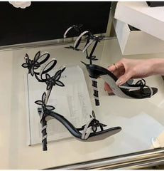 Black high-heeled sandals with floral designs on a reflective surface.