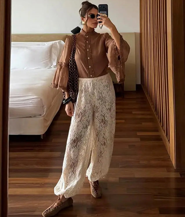 Woman taking a mirror selfie wearing a brown blouse and white lace pants in a bedroom.
