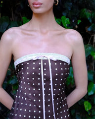 Woman wearing a strapless dress with polka dot pattern and lace details, standing against a blurred green leafy background.