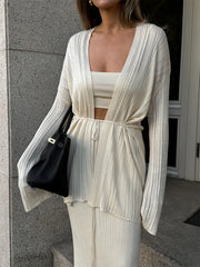 Woman wearing a white outfit with a black handbag against a gray wall.