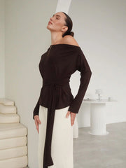 Woman wearing a black off-shoulder top and white pants in a minimalistic room.