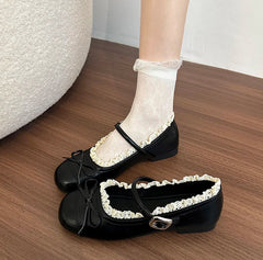 A pair of black ballet flats with a decorative butterfly knot and lace trim, featuring a side buckle strap.