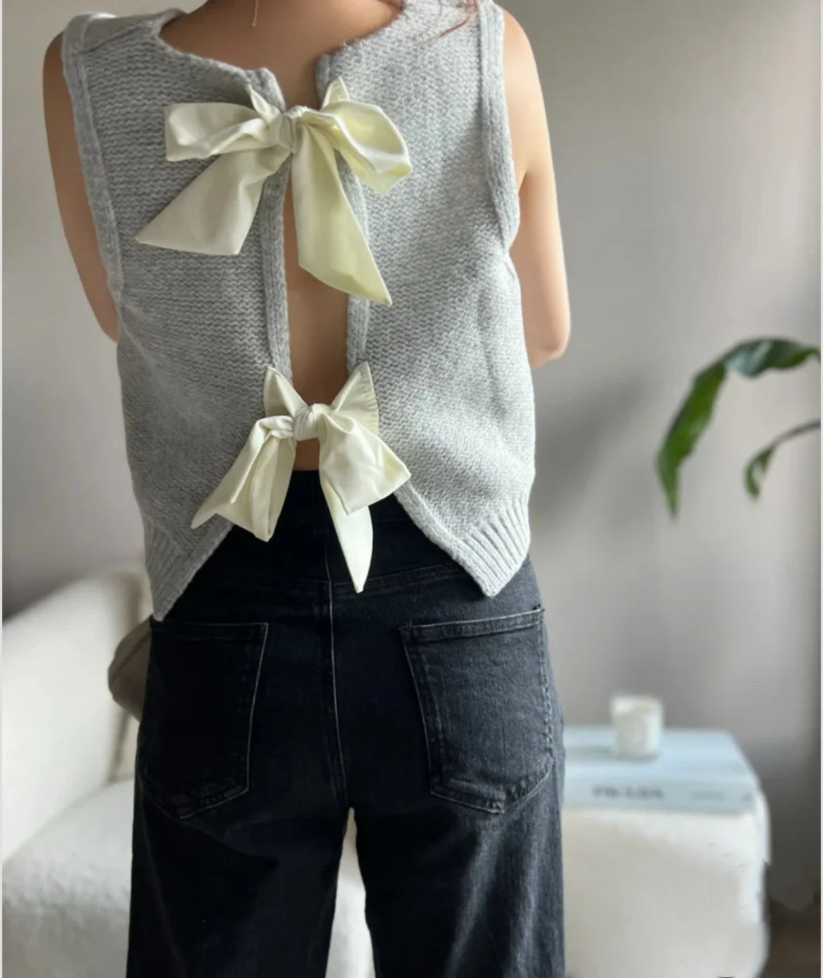 Person wearing a gray sleeveless top with white bows and dark jeans in an indoor setting.