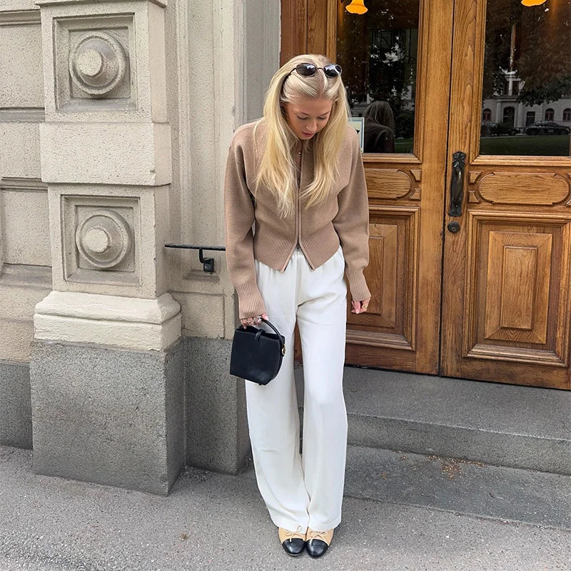 Woman in beige jacket and white pants standing outside a building with wooden doors.