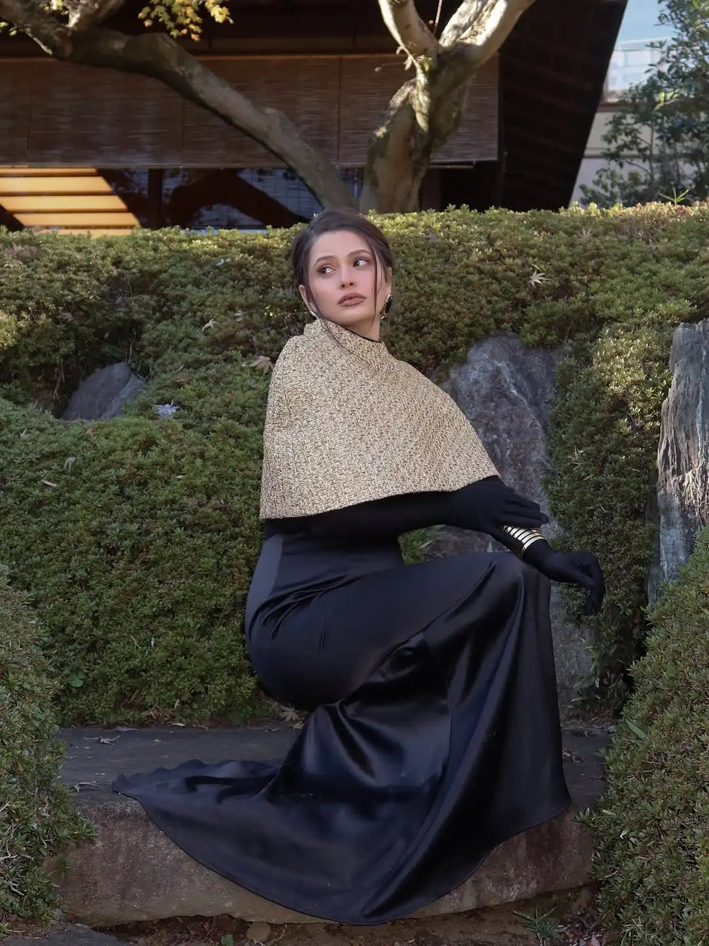 Woman in a black dress with a beige shawl sitting on a stone in a garden setting.