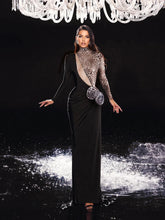 Woman in a black evening gown with a textured shawl and clutch, standing under a chandelier.