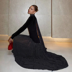 Woman in a black and white polka dot dress holding a red clutch against a neutral wall.
