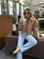 A woman sitting outdoors holding a cup, wearing sunglasses and a khaki jacket.