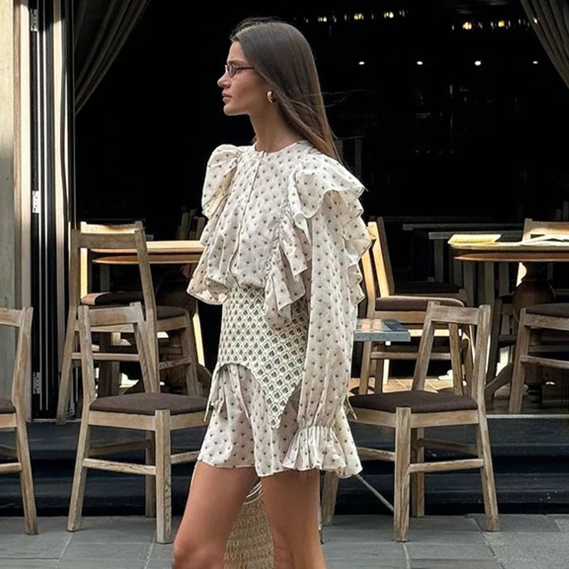 Woman in a white patterned dress standing in an outdoor setting with tables and chairs.