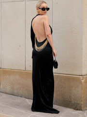 Woman wearing a black evening gown with a gold necklace against a beige wall.
