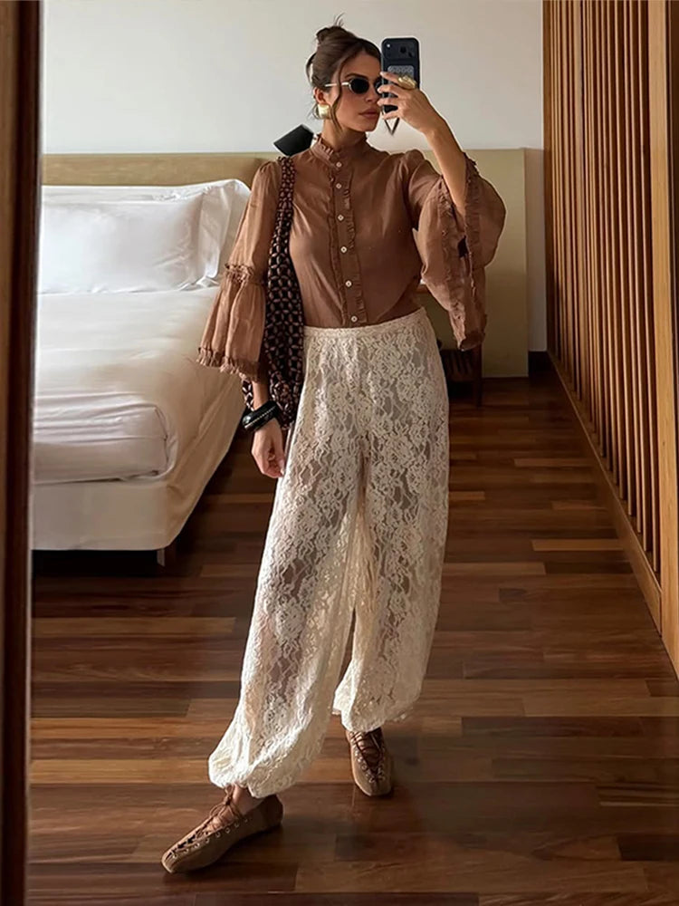 Woman taking a mirror selfie wearing a brown blouse and white lace pants in a bedroom.