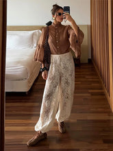 Woman taking a mirror selfie wearing a brown blouse and white lace pants in a bedroom.