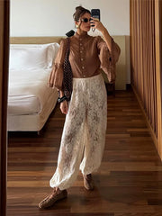 Woman taking a mirror selfie wearing a brown blouse and white lace pants in a bedroom.