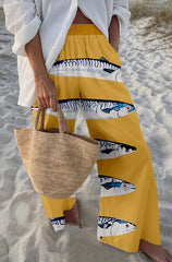 Person wearing yellow pants with fish pattern and holding a straw bag on sandy ground