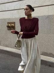 Woman in burgundy top and white pants standing against a gray wall.