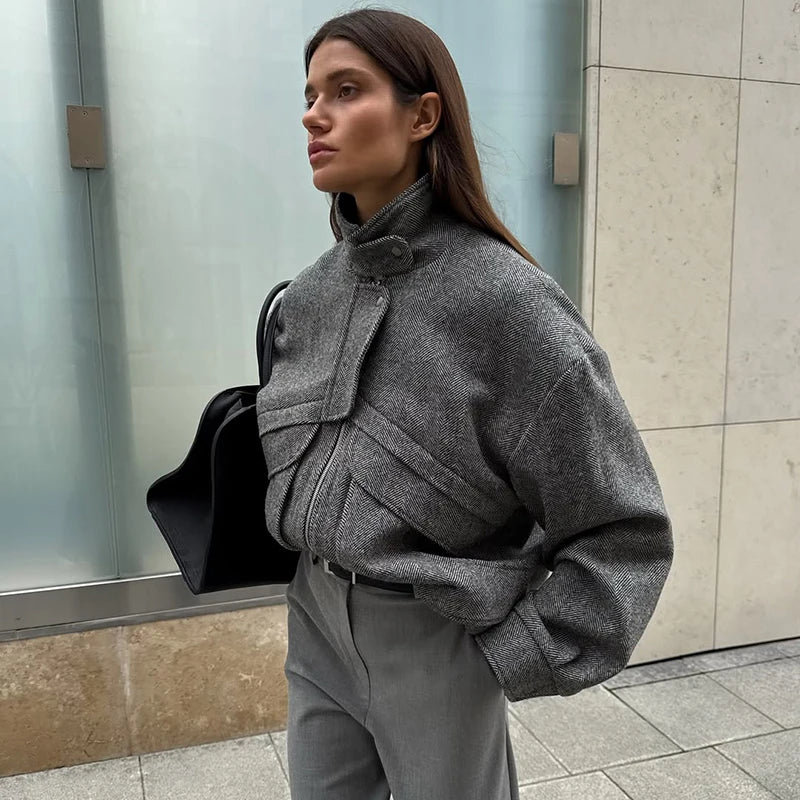 Woman wearing a gray coat and holding a black bag against a tiled wall.
