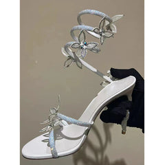 White high-heeled sandal with decorative straps and flowers held by a gloved hand against a neutral background