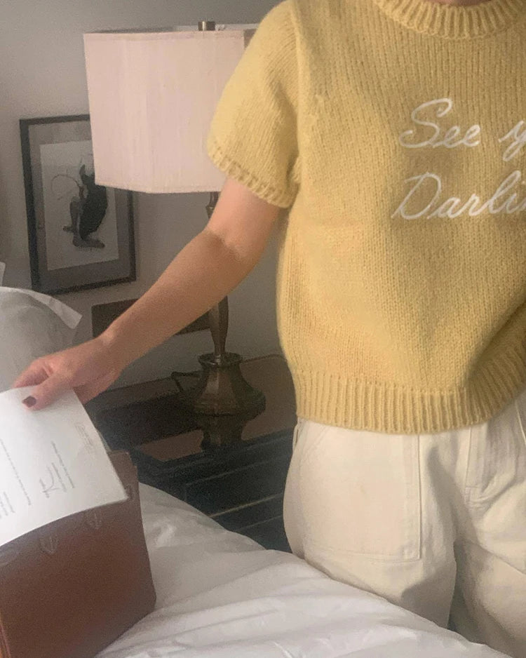 Person wearing a yellow sweater with text, sitting on a bed in a bedroom.