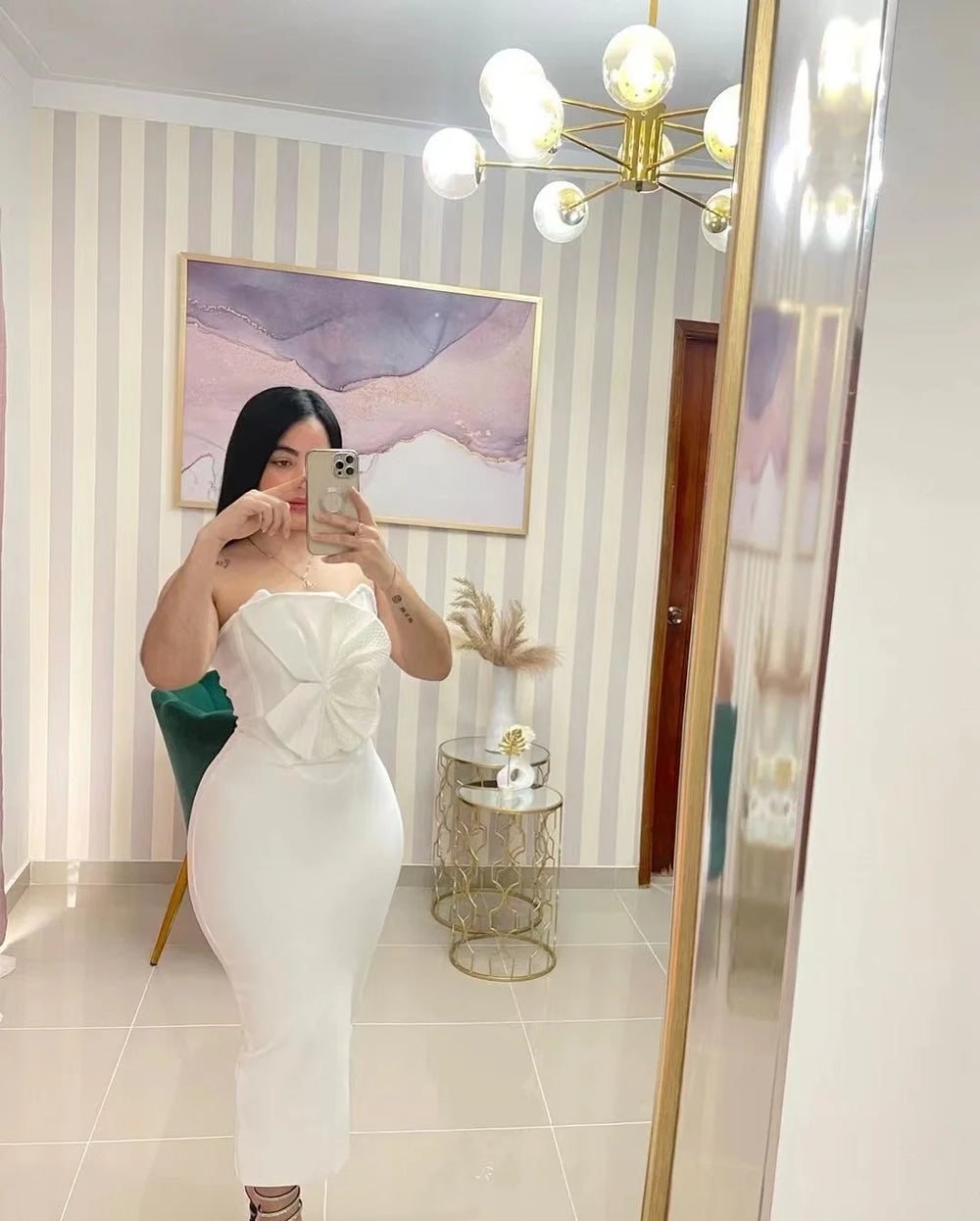 Person taking a mirror selfie wearing a white strapless dress in a room with striped wallpaper and a chandelier.