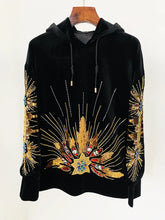 Black velvet hoodie with gold sequin and beaded sunburst embellishment, displayed on a hanger.