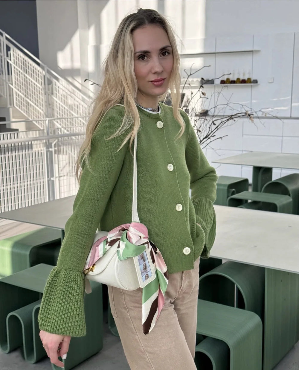 Woman in a green cardigan holding a colorful handbag in an outdoor setting.