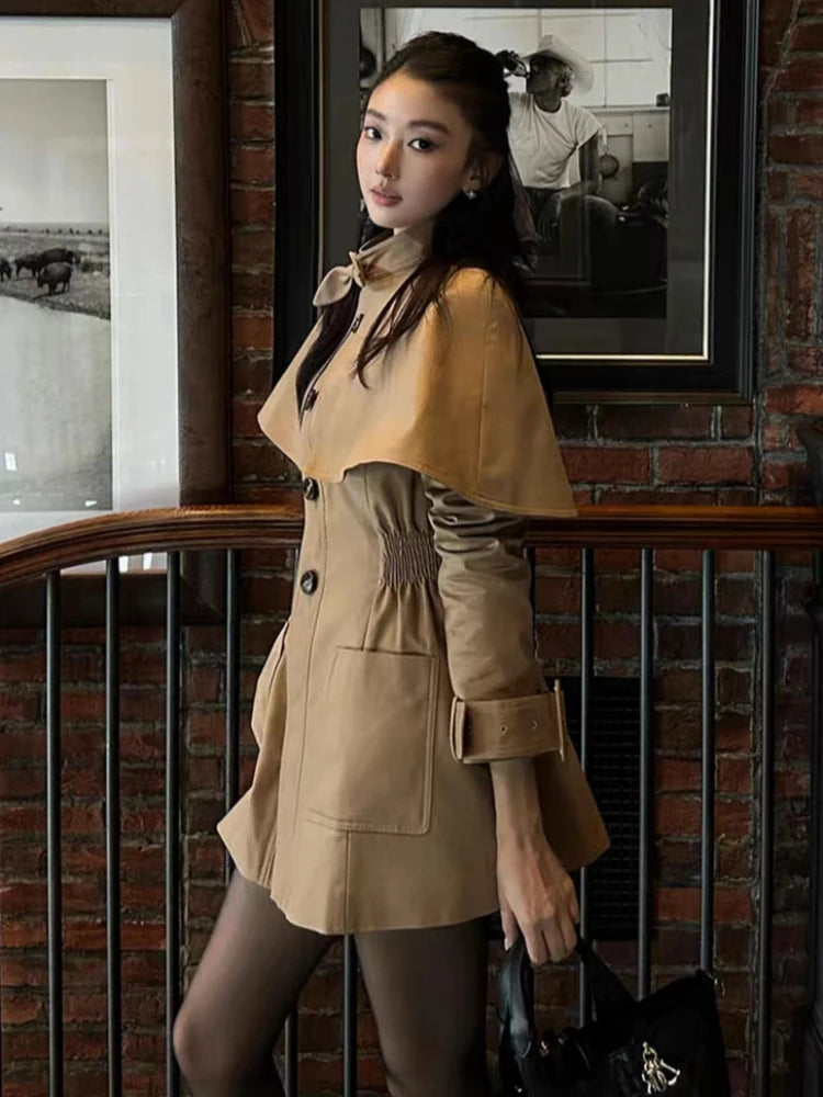 Woman in a beige trench coat standing in a room with brick walls and framed pictures.