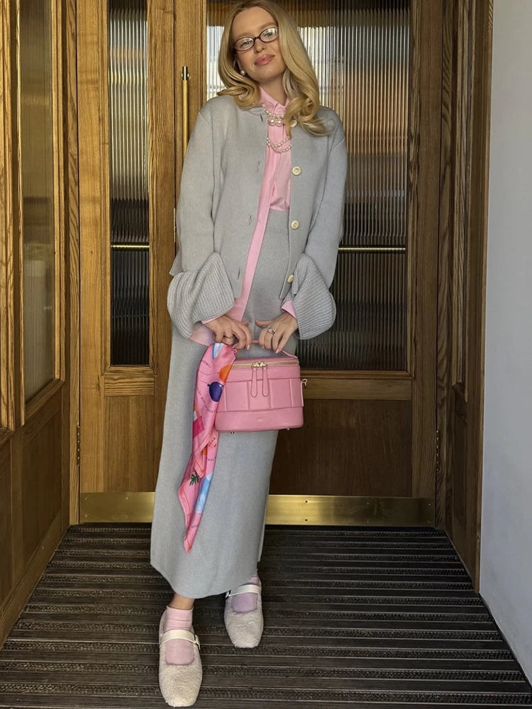 Woman in gray coat and pink shoes holding a pink handbag in front of a wooden door.