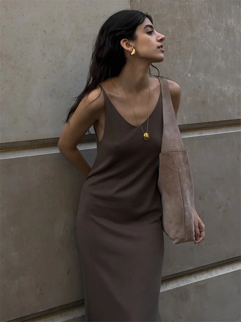Woman in a brown dress holding a beige bag against a neutral wall.