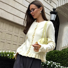 A woman wearing a white cardigan and black pants, holding a yellow handbag, standing outdoors.