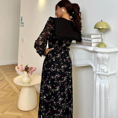 Woman in a black floral dress standing in a room with a white fireplace and decorative items.