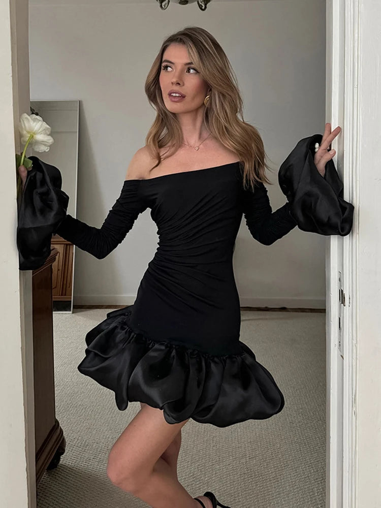 Woman wearing a black off-shoulder dress with large gloves indoors.