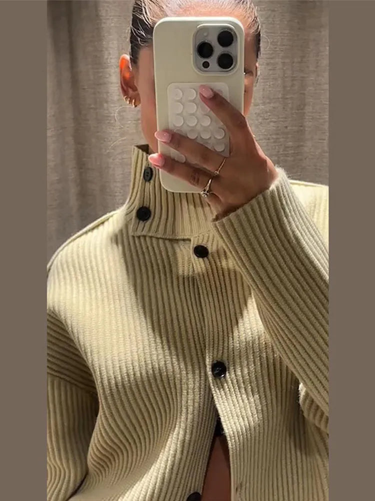 Person wearing a beige ribbed sweater with black buttons, holding a phone with a white case.